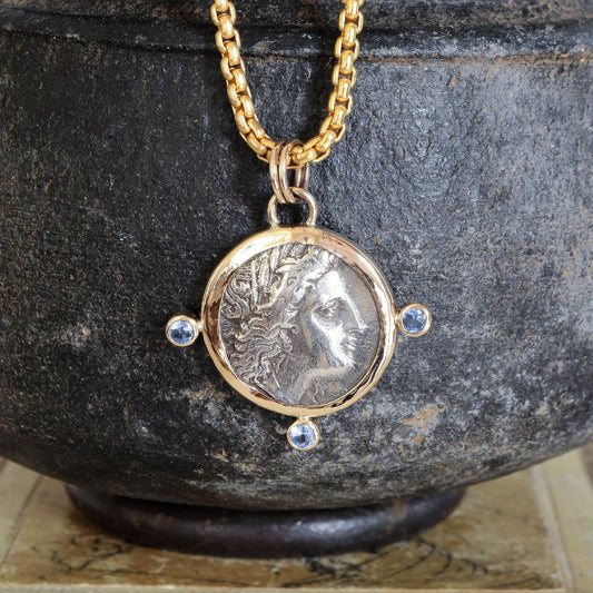 Necklace with a pendant featuring an ancient coin with Tanzanite gemstones, displayed against a dark stone background.