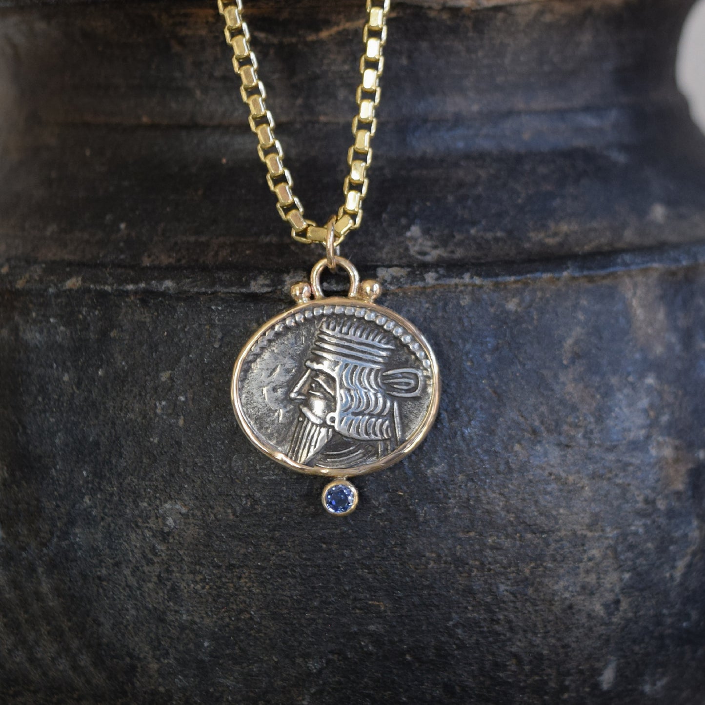 Gold necklace with an ancient coin pendant with sapphire featuring an emblem on a dark stone surface