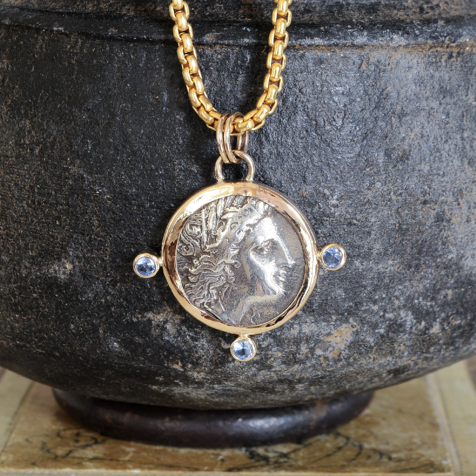 Necklace with a pendant featuring an ancient coin with Tanzanite gemstones, displayed against a dark stone background.